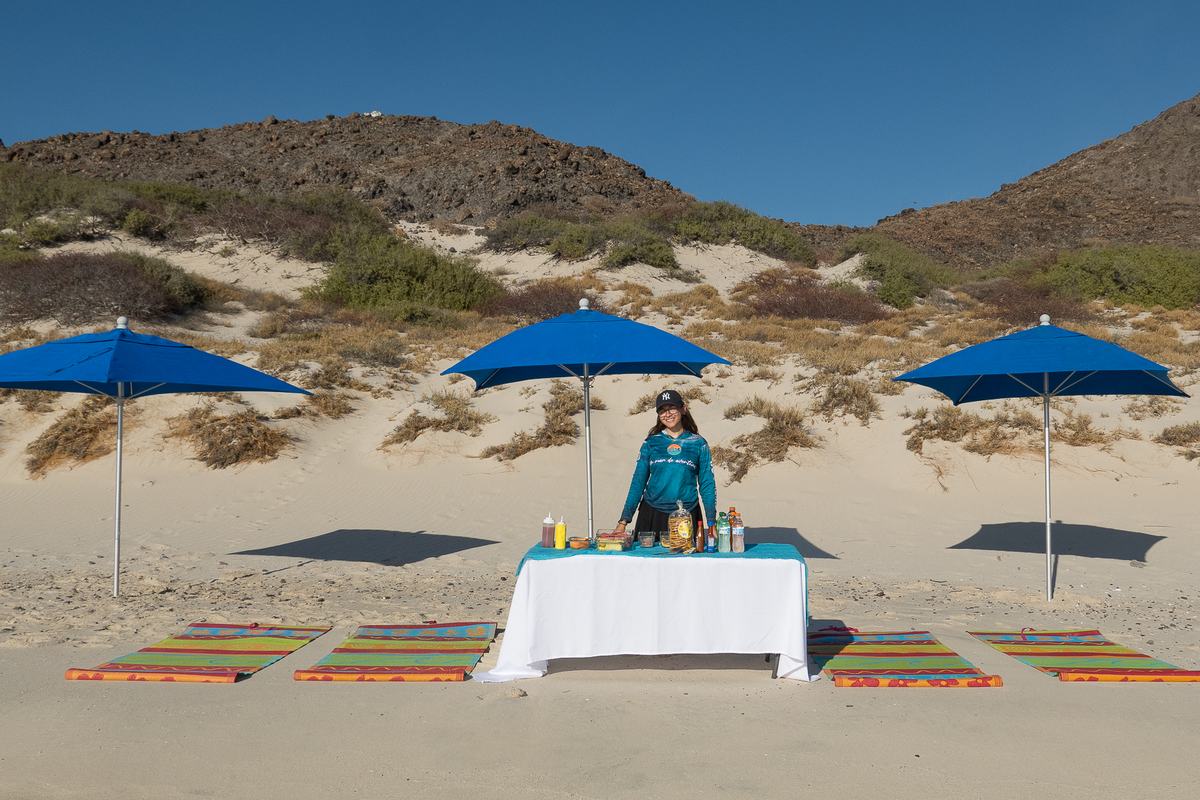 Montaje de Pita’s Tours en la playa: sombrillas azules, petates y la mesa con la comida servida